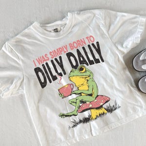 May include: White t-shirt featuring a green frog graphic, holding a teacup, and sitting on a mushroom. The text reads "I WAS SIMPLY BORN TO DILLY DALLY". A pair of white and grey trainers with black stripes are next to the shirt.
