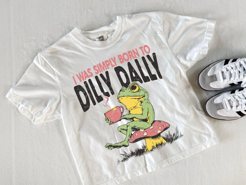 May include: White t-shirt featuring a green frog graphic, holding a teacup, and sitting on a mushroom. The text reads "I WAS SIMPLY BORN TO DILLY DALLY". A pair of white and grey trainers with black stripes are next to the shirt.