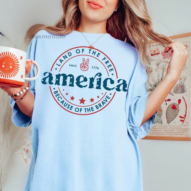 May include: Light blue t-shirt with the word "america" in a circle design, with the text "LAND OF THE FREE SINCE 1776 BECAUSE OF THE BRAVE". A white mug with a sun design is held in the left hand.