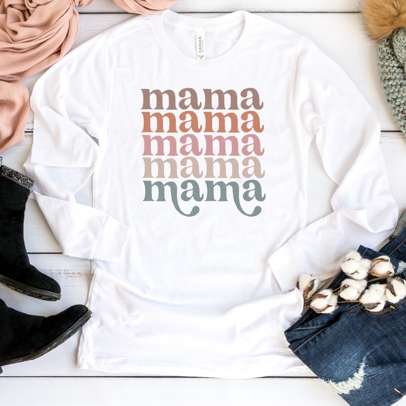 Puede incluir: Camiseta blanca de manga larga con el texto "mama" repetido cinco veces en un dise&ntilde;o apilado. Cada "mama" es de un color diferente, comenzando con un rosa claro y pasando a un azul claro.