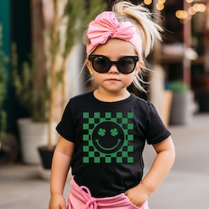 May include: A young child wearing a black t-shirt with a green and black checkered pattern. The shirt features a smiley face with shamrock eyes. The child is also wearing a pink bow headband and black sunglasses.