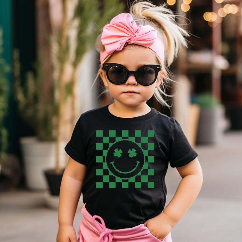 May include: A young child wearing a black t-shirt with a green and black checkered pattern. The shirt features a smiley face with shamrock eyes. The child is also wearing a pink bow headband and black sunglasses.