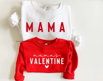 Pode incluir: Duas sweatshirts com tema do Dia dos Namorados em vermelho e branco. A maior sweatshirt é branca com a palavra "MAMA" em letras vermelhas. A menor sweatshirt é vermelha com o texto "mama's VALENTINE" em letras brancas e um pequeno coração branco.