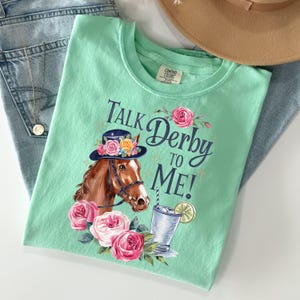 Może przedstawiać: Miętowa koszulka z napisem "Talk Derby to Me!" i grafiką konia w kapeluszu z kwiatami. Projekt zawiera r&oacute;żowe r&oacute;że i koktajl.