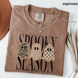 May include: An espresso-colored t-shirt featuring the text "SPOOKY SEASON" in black capital letters. Three ghost-shaped graphics are centered above the text, each with a different pattern: camouflage, leopard print, and a tan and brown checkerboard.