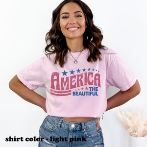 Pu&ograve; includere: Una maglietta rosa chiaro con una grafica in stile vintage che dice "America The Beautiful" in rosso, bianco e blu. La grafica presenta stelle e strisce.