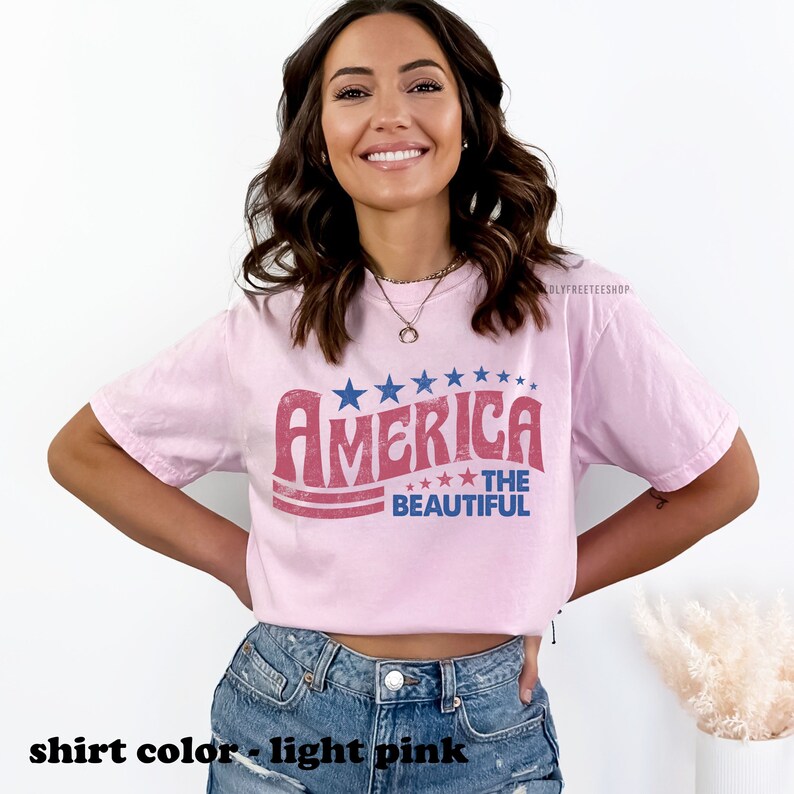 Pu&ograve; includere: Una maglietta rosa chiaro con una grafica in stile vintage che dice "America The Beautiful" in rosso, bianco e blu. La grafica presenta stelle e strisce.