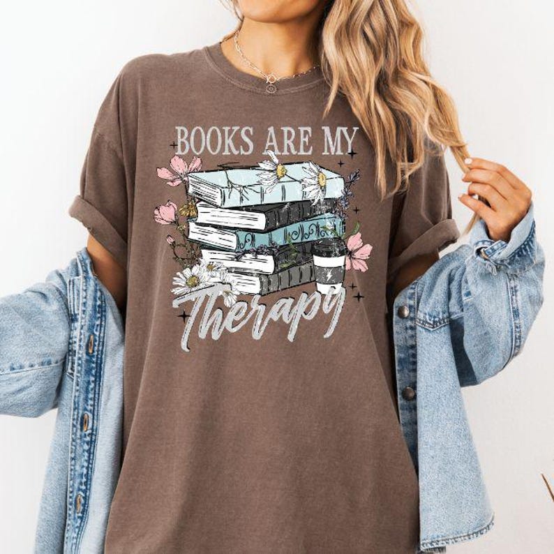 May include: Brown t-shirt with the text "BOOKS ARE MY Therapy" and a graphic of stacked books, flowers, and a coffee cup. The design features light blue, black, and white elements.