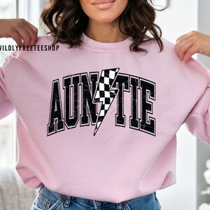 K&ouml;nnte beinhalten: Ein hellrosa Sweatshirt mit Rundhalsausschnitt und dem Wort AUNTIE in schwarzer College-Schrift mit abgenutzter Textur. Der Buchstabe I ist durch einen schwarz-wei&szlig; karierten Blitz ersetzt. Der Text @WILDLYFREETEESHOP erscheint in der Ecke.