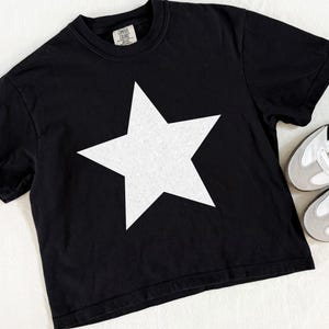 May include: Black t-shirt with a large white star graphic. The shirt is paired with white and gray sneakers with black stripes. The t-shirt has a crew neck and short sleeves. The image is taken from an overhead angle.