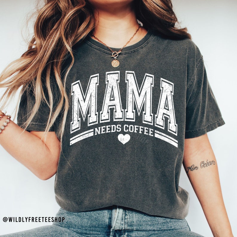May include: A charcoal grey short-sleeved t-shirt featuring white distressed lettering that reads MAMA NEEDS COFFEE with a small heart icon centred below the text. The shirt has a crew neckline and a relaxed fit, showcasing a vintage-inspired graphic.
