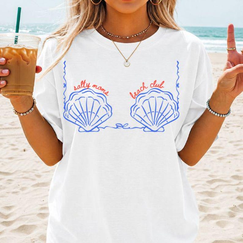 May include: White t-shirt with a blue seashell design and the text "salty moms beach club" in red. The seashells are drawn with blue outlines and lines. The shirt is a casual, beach-themed apparel item.