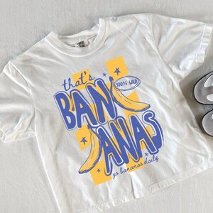 Pu&ograve; includere: T-shirt bianco con grafica blu e gialla con la scritta "that's BANANAS" con illustrazioni di banane. La maglietta ha anche il testo "go bananas daily" e "100% WILD". Un paio di scarpe da ginnastica bianche e grigie con strisce nere sono accanto alla maglietta.