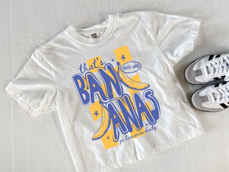 Pu&ograve; includere: T-shirt bianco con grafica blu e gialla con la scritta "that's BANANAS" con illustrazioni di banane. La maglietta ha anche il testo "go bananas daily" e "100% WILD". Un paio di scarpe da ginnastica bianche e grigie con strisce nere sono accanto alla maglietta.