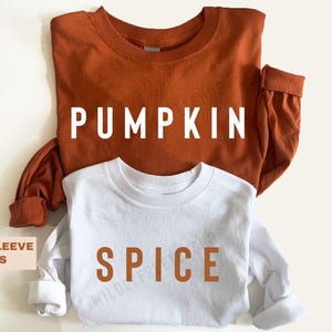 May include: A rust-coloured long-sleeve shirt with the word PUMPKIN printed in white letters and a white long-sleeve shirt with the word SPICE printed in rust-coloured letters. Both shirts feature a crew neckline and long sleeves with cuffs.