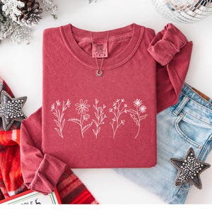 May include: A maroon long-sleeve shirt with a white floral design. The shirt has a crew neck and rolled-up sleeves. A silver necklace with a sun pendant is on the shirt. The shirt is paired with blue jeans and decorative stars.