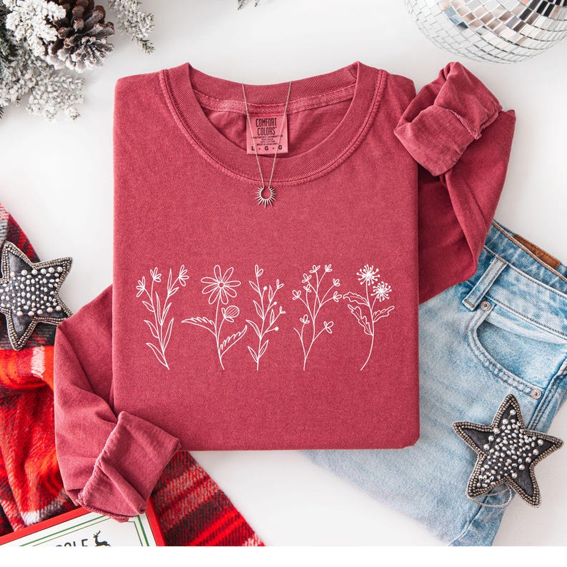 May include: A maroon long-sleeve shirt with a white floral design. The shirt has a crew neck and rolled-up sleeves. A silver necklace with a sun pendant is on the shirt. The shirt is paired with blue jeans and decorative stars.