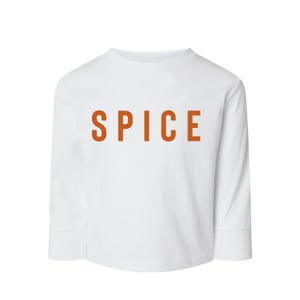 May include: A white long-sleeved shirt featuring the word SPICE printed in bold, orange, sans-serif capital letters across the centre of the chest. The garment has a crew neckline and straight sleeves with ribbed cuffs, providing a clean look.