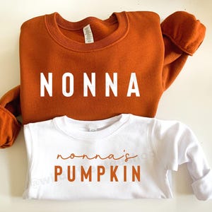 May include: Two sweatshirts, one burnt orange and one white. The orange sweatshirt has the word "NONNA" in white block letters. The white sweatshirt has the words "nonna's PUMPKIN" in orange script and block letters.