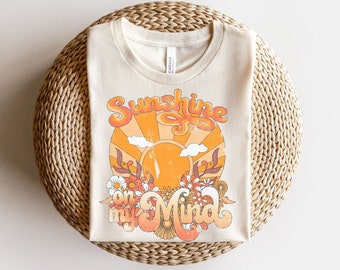 May include: A beige t-shirt with a retro-style graphic design featuring a sun with rays, clouds, and flowers. The text "Sunshine on my Mind" is written in a groovy font.