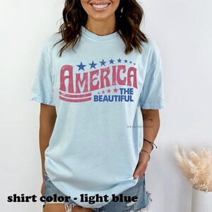 Pu&ograve; includere: Una maglietta blu chiaro con una grafica in stile vintage che dice "America The Beautiful" in lettere rosse e blu con stelle.