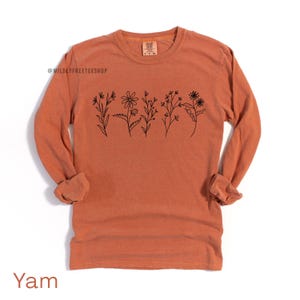 May include: A long-sleeved, orange-colored shirt with a crew neck. The shirt features a black line drawing of various flowers across the chest. The text "Yam" is at the bottom of the shirt.