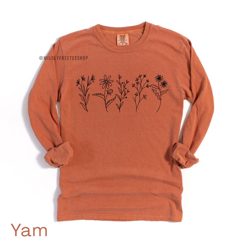 May include: A long-sleeved, orange-colored shirt with a crew neck. The shirt features a black line drawing of various flowers across the chest. The text "Yam" is at the bottom of the shirt.