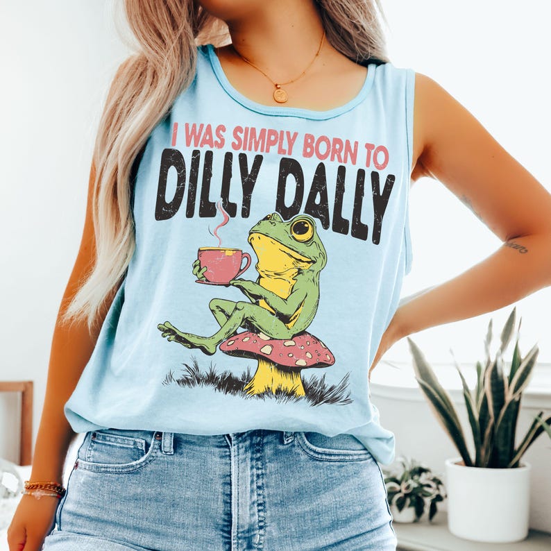 Op de afbeelding: Lichtblauwe tanktop met de tekst "I WAS SIMPLY BORN TO DILLY DALLY". De afbeelding toont een groene kikker die op een paddenstoel zit en een roze kopje vasthoudt. De kikker drinkt uit het kopje. De tanktop is gemaakt van zacht materiaal.