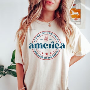 May include: Cream-colored t-shirt with a patriotic design. The design includes the word "America" in blue, surrounded by a red circle with the text "Land of the Free" and "Because of the Brave." A peace sign is also included.