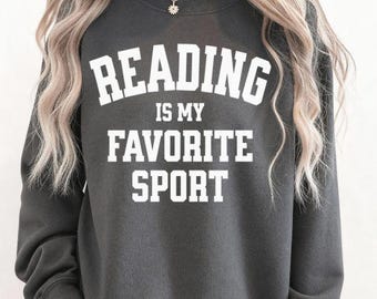 May include: Dark grey sweatshirt with the white text "READING IS MY FAVORITE SPORT". The sweatshirt has a crew neck and long sleeves. The image shows the upper body of a person wearing the sweatshirt.