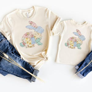 May include: Two cream-coloured t-shirts, one adult and one child size, each with a patchwork bunny design. The bunny is made of floral and gingham fabrics. Dark blue jeans and dried grass complete the Easter theme.