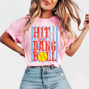 May include: Pale pink t-shirt featuring the text "HIT the DANG BALL" in red and yellow, with a softball graphic. The text is set against a blue and white striped background.