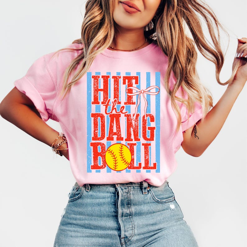 May include: Pale pink t-shirt featuring the text "HIT the DANG BALL" in red and yellow, with a softball graphic. The text is set against a blue and white striped background.