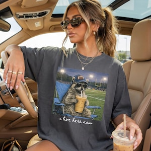 Op de afbeelding: Donkergrijs t-shirt met een wasbeer met een zonnebril en een footballpet, die een drankje vasthoudt en in een blauwe stoel zit. De afbeelding toont een voetbalveld met de tekst "i live here now".