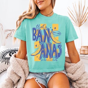 Pu&ograve; includere: T-shirt turchese con la scritta "that's BANANAS" in blu, con illustrazioni di banane e il testo "100% WILD" e "go bananas daily". La maglietta ha un girocollo e maniche corte.