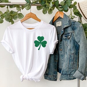 May include: White t-shirt with a green glitter shamrock on the front. The shirt is hanging on a hanger with a denim jacket next to it.
