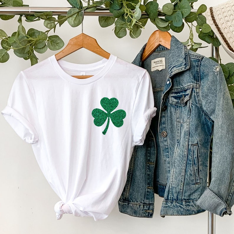 May include: White t-shirt with a green glitter shamrock on the front. The shirt is hanging on a hanger with a denim jacket next to it.