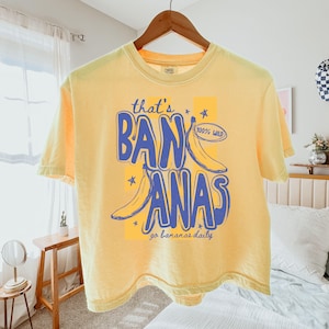 Pu&ograve; includere: T-shirt giallo chiaro con la scritta blu "that's BANANAS", illustrazioni di banane e la scritta "100% WILD". La maglietta &egrave; appesa a una gruccia di legno.