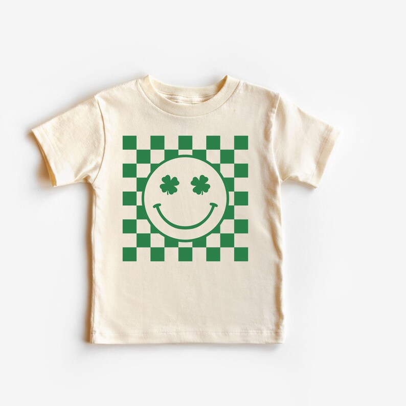 May include: A cream-colored t-shirt featuring a green and white checkered pattern. A smiley face with shamrock eyes is centered on the shirt. The shirt is made of a soft material and is suitable for children.