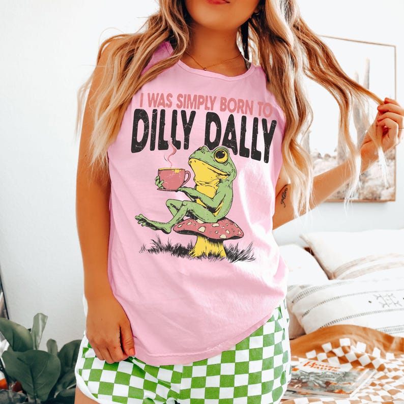 Op de afbeelding: Lichtroze T-shirt met de tekst "I WAS SIMPLY BORN TO DILLY DALLY" en een afbeelding van een groene kikker op een paddenstoel, met een kopje koffie. De kikker is geel en groen.