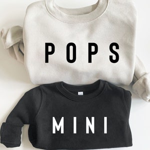 Könnte beinhalten: Zwei Sweatshirts, eines beige und eines schwarz, mit den Worten "POPS" und "MINI" in Schwarz und Weiß bedruckt.
