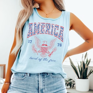 May include: Light blue tank top with the word "AMERICA" in red and blue block letters. Below the word "AMERICA" is a graphic of an eagle with the numbers "17" and "76" on either side. The words "land of the free" are written below the eagle.