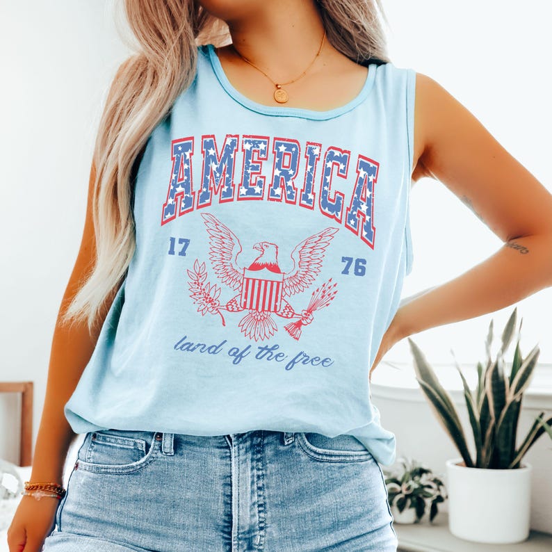 May include: Light blue tank top with the word "AMERICA" in red and blue block letters. Below the word "AMERICA" is an eagle graphic with the numbers "17" and "76" on either side. The words "land of the free" are printed below the eagle.