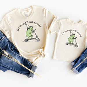 Puede incluir: Dos camisetas color crema, una para adultos y otra para niños, con un caprichoso gráfico de una rana montada en un scooter y el texto "Off to commit dad foolery" y "Off to commit mini foolery". Vaqueros azules junto a las camisetas.