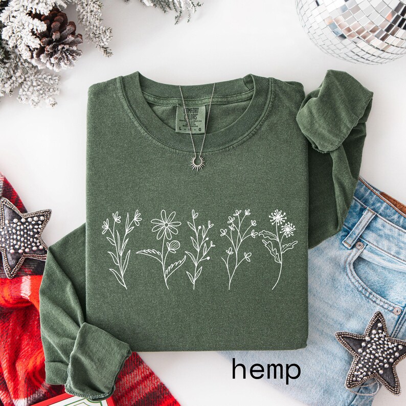 May include: A dark green long-sleeved shirt with a white floral design and the word "hemp" at the bottom. A silver necklace with a sun pendant is on the shirt. The shirt is folded on a white surface with denim jeans and holiday decorations.