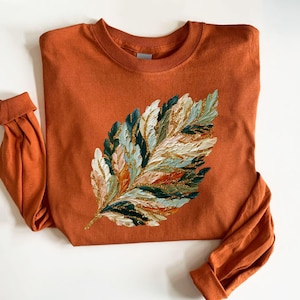 Op de afbeelding: Roestkleurig sweatshirt met lange mouwen met een gestructureerd bladontwerp. Het blad is beschilderd met wit, groen, goud en perzik. De manchetten zijn opgevouwen. Het ontwerp geeft de trui een artistieke uitstraling.