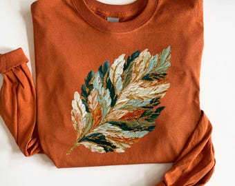 Puede incluir: Una camiseta de manga larga con cuello redondo en un color naranja quemado sólido. El centro de la camiseta presenta un gráfico grande y texturizado de una pluma en tonos verde oscuro, crema y naranja óxido con detalles de purpurina dorada.