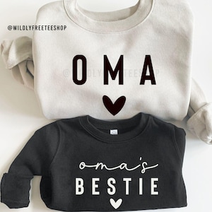 Puede incluir: Dos sudaderas, una blanca con texto negro que dice "OMA" y un corazón negro, y una negra con texto blanco que dice "oma's BESTIE" y un corazón blanco.