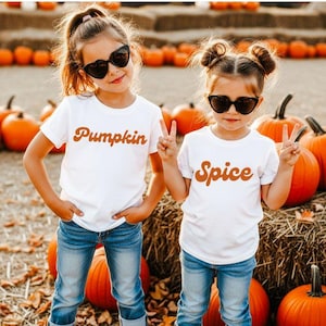 May include: Two white short-sleeved t-shirts featuring orange retro-style text. One shirt displays the word Pumpkin and the other displays the word Spice. Both shirts are paired with blue denim jeans and black heart-shaped sunglasses.