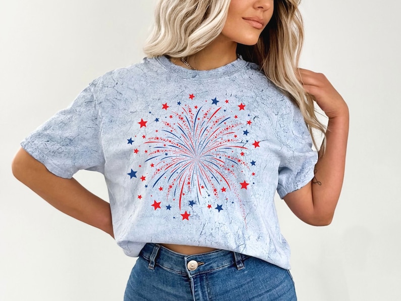 Op de afbeelding: Een lichtblauw T-shirt met een rood, wit en blauw vuurwerkmotief. Het vuurwerk is omringd door rode en blauwe sterren.
